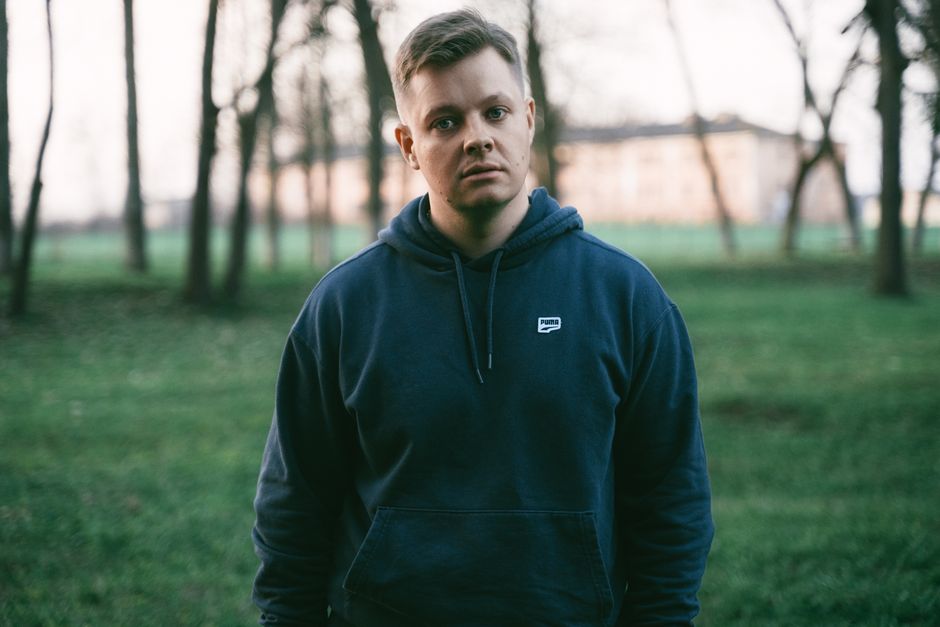 A man in a hoodie stands in a grassy park with trees in the background, captured during daytime.