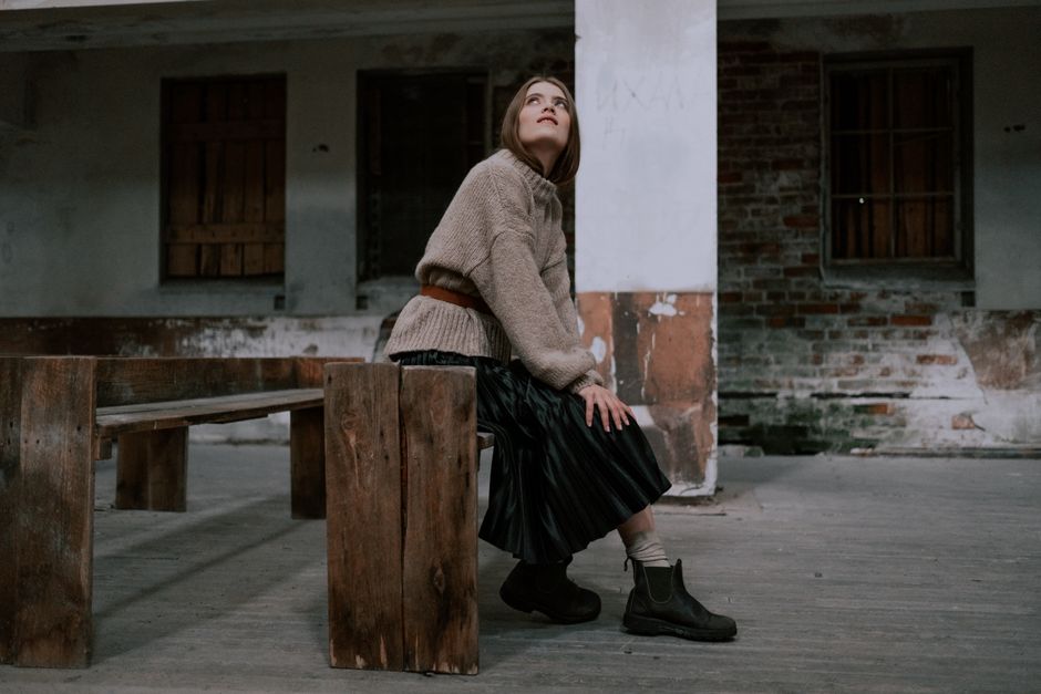 A woman in a brown sweater and black skirt sits on a wooden bench in a rustic setting, looking contemplative.
