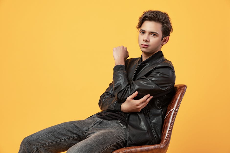 Unemotional young handsome male with short dark hair in leather coat and jeans looking at camera while chilling on chair with arms crossed