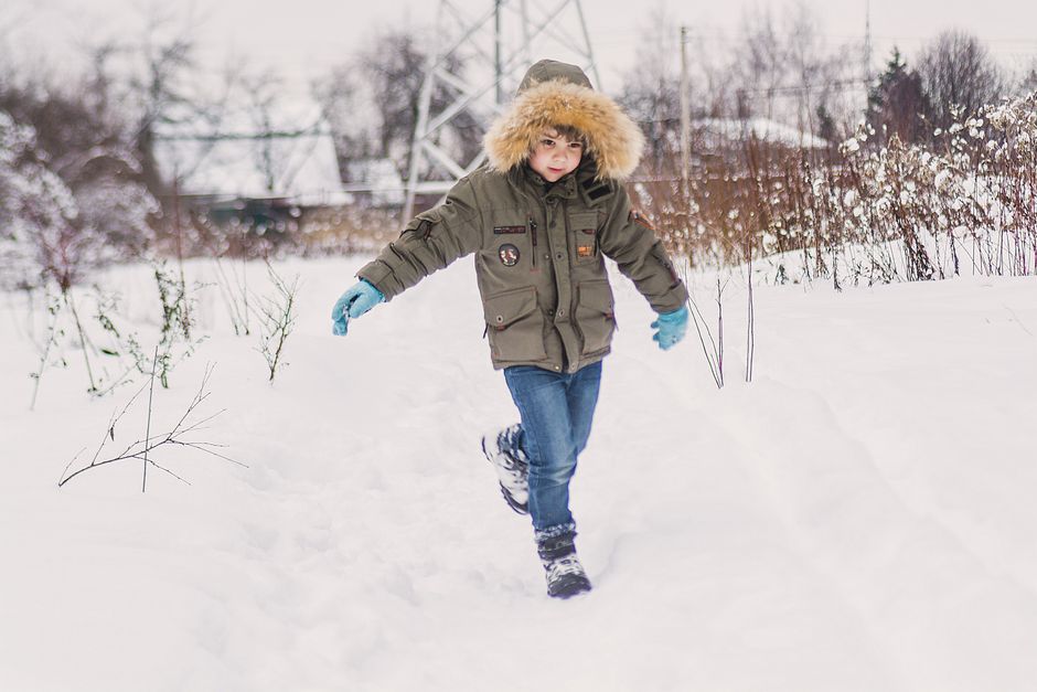 Young boy running through a snowy winter landscape, wearing a cozy fur-lined parka and gloves.