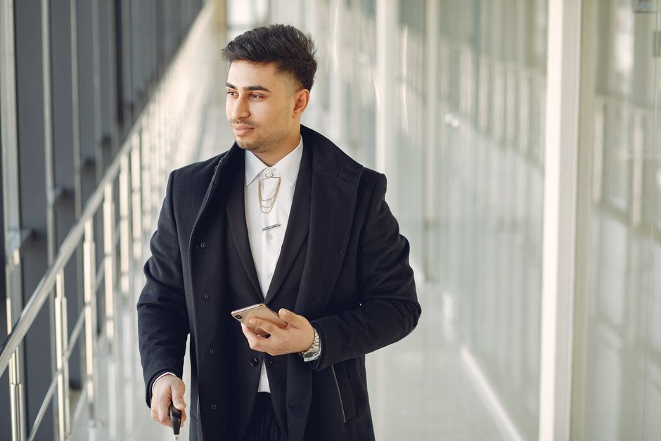 Thoughtful ethnic male in formal suit standing in modern hallway and holding mobile phone in hand while looking through window