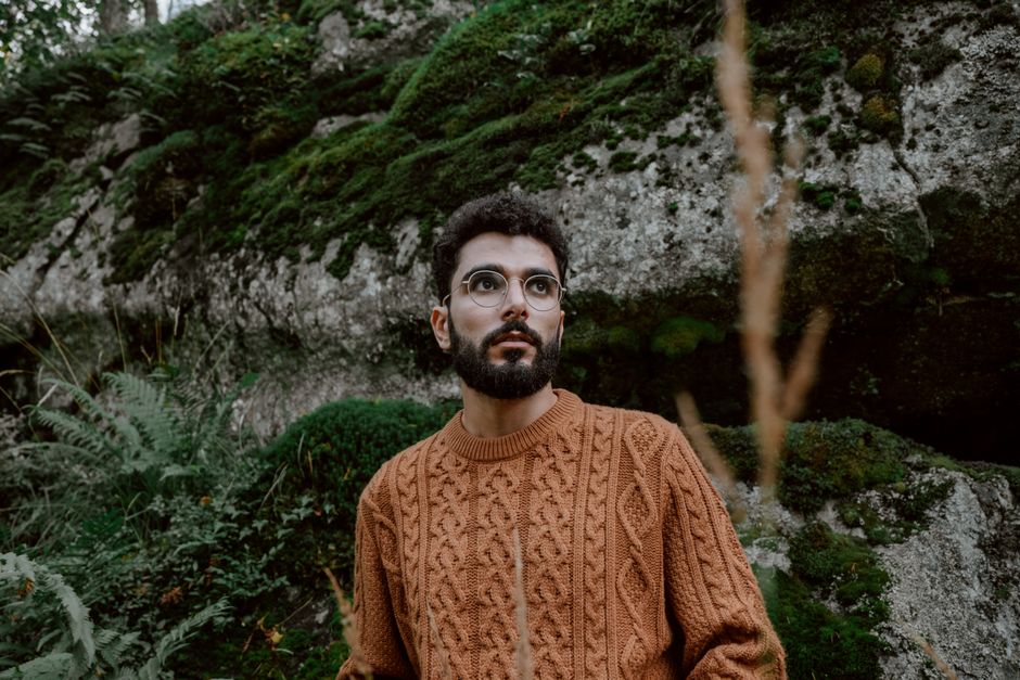 Young man in glasses and a knitted sweater in lush outdoor setting.