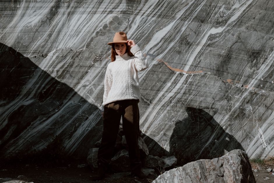 Stylish woman posing in a fedora hat and sweater against a dramatic marble backdrop outdoors.
