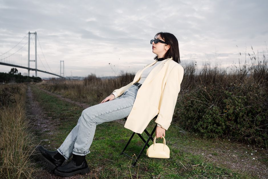 A stylish woman in sunglasses relaxes on a chair by a bridge in nature.