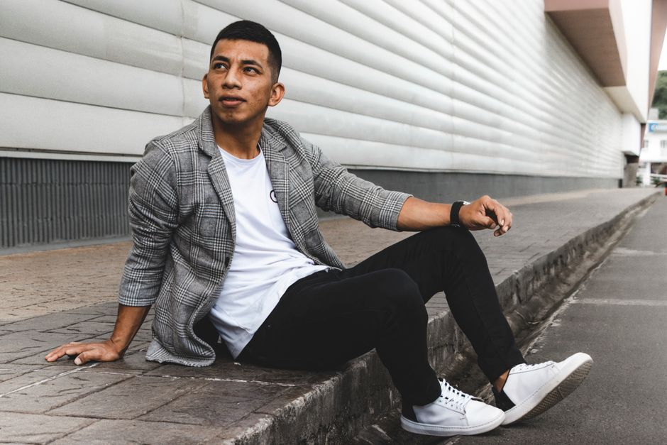 Stylish man sitting on the pavement in a modern urban setting wearing a plaid blazer and sneakers.