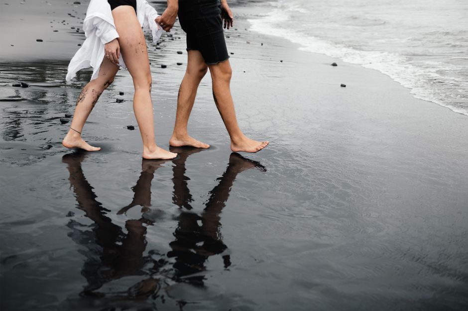 A barefoot couple walking along a black sand beach, reflecting off wet sand.