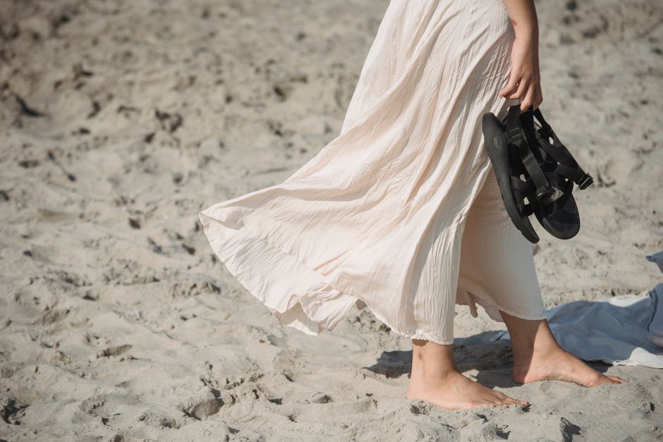 A woman strolls barefoot on a beach, holding her sandals with a breeze in her dress.