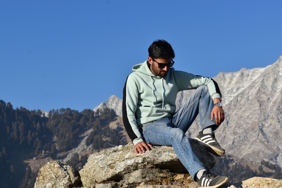 A man in casual attire sitting on rocks with mountains in the background under clear blue skies.
