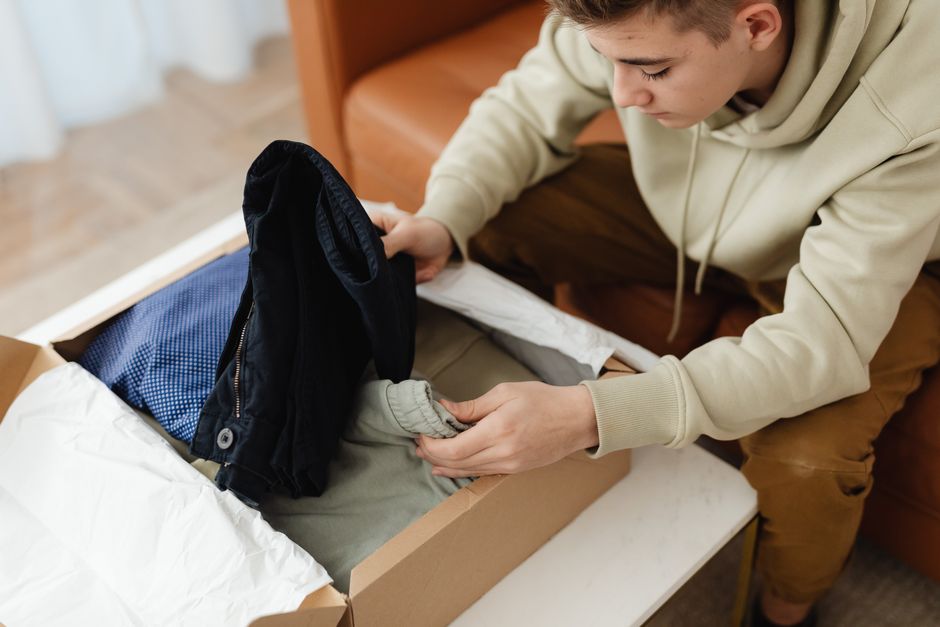 Young man opening a box with clothes on a sofa. Casual, comfortable setting indoors.