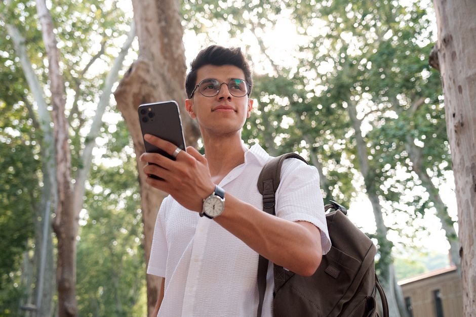 Confident young adult with backpack and smartphone in a sunny park setting, embracing modern lifestyle.
