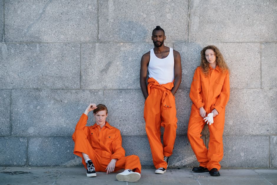 Three adults in orange uniforms pose against a stone wall, showcasing diversity.