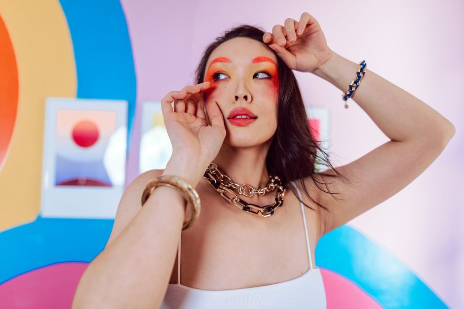 A young woman poses confidently with vibrant makeup, set against a colorful, artistic background.