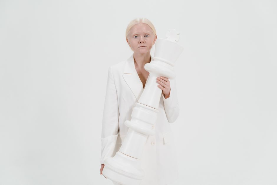 Woman in white outfit holding a large white chess king against a plain background.