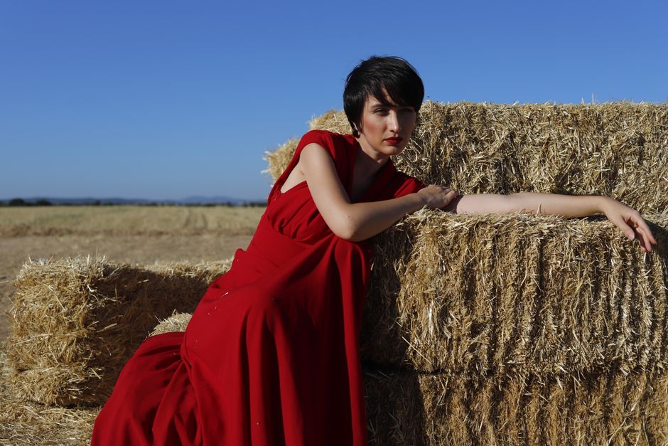 Fashion model in a red dress posing elegantly on hay bales in a rural field.