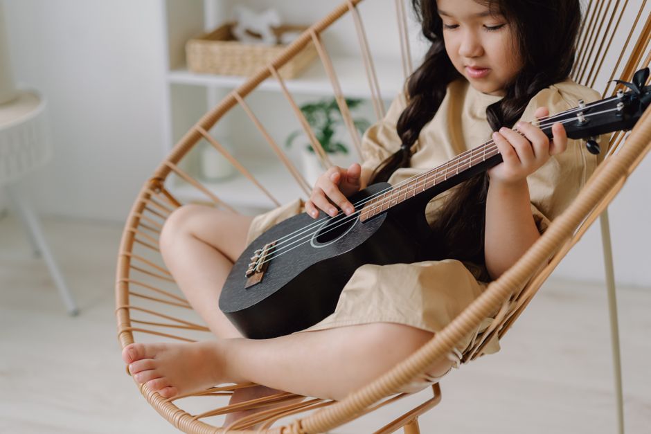 Little girl sitting on a rattan chair playing a ukulele indoors.