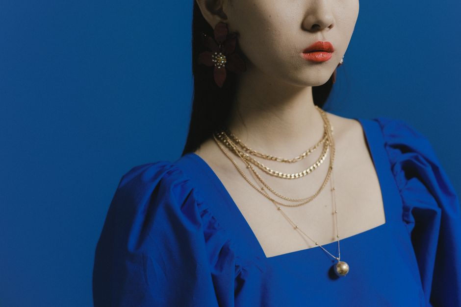 Portrait of a woman in a blue dress wearing gold necklaces, set against a blue background.