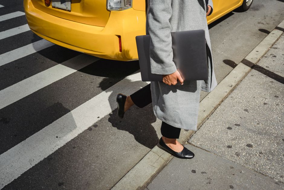 Crop faceless female in trendy coat holding laptop while crossing asphalt road near yellow taxi in city