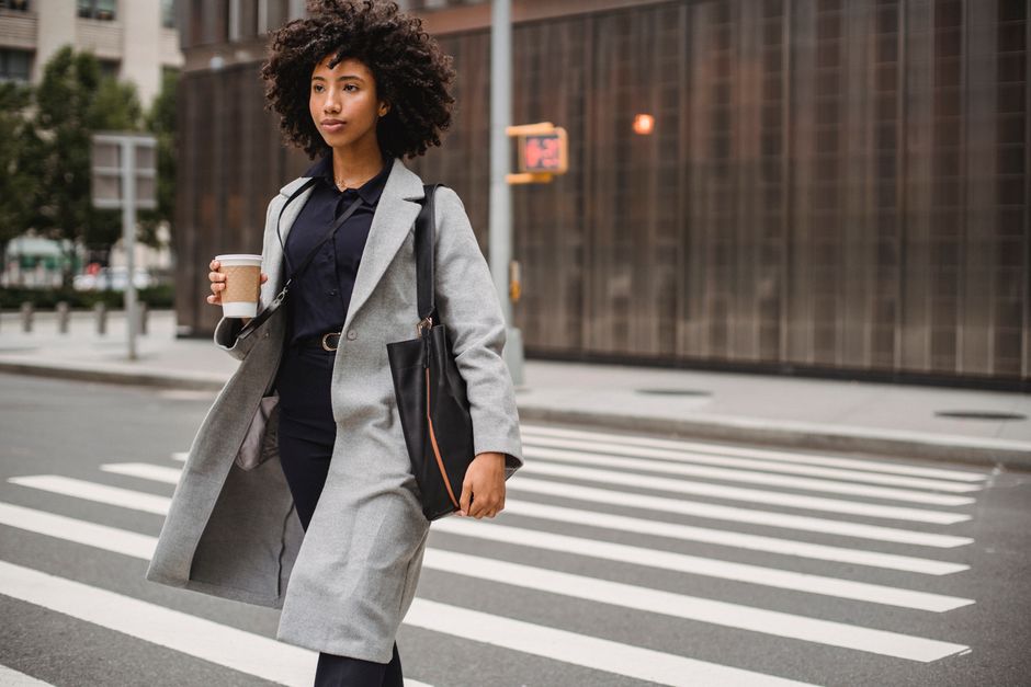 Young woman crossing an urban street holding coffee, exuding confidence in a gray coat.