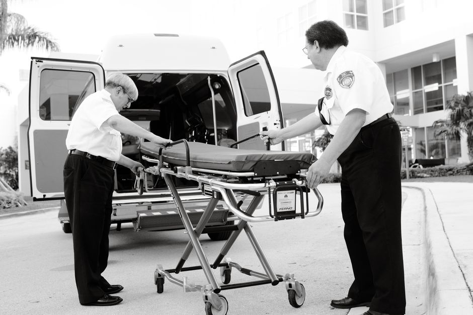 Two paramedics carefully load a stretcher into an ambulance outside a facility.