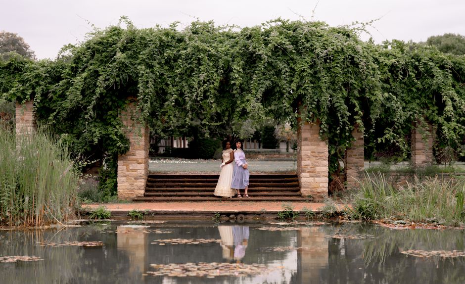 Two elegantly dressed women stand in a lush garden pergola by a reflective pond.