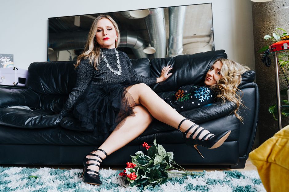 Stylish women in glamorous outfits posing on a black leather sofa indoors.