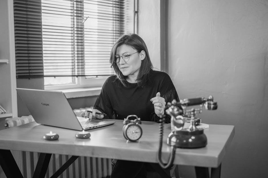 Black and white photo of a woman working at a desk with a laptop and vintage telephone.