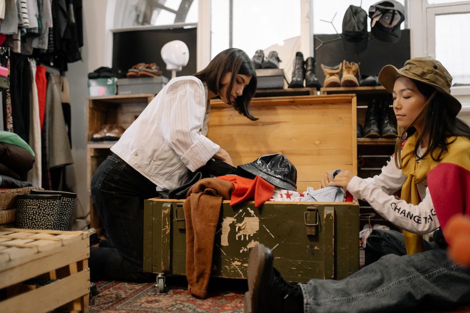 Two women sorting through vintage clothing in a thrift shop, surrounded by shoes and hats.