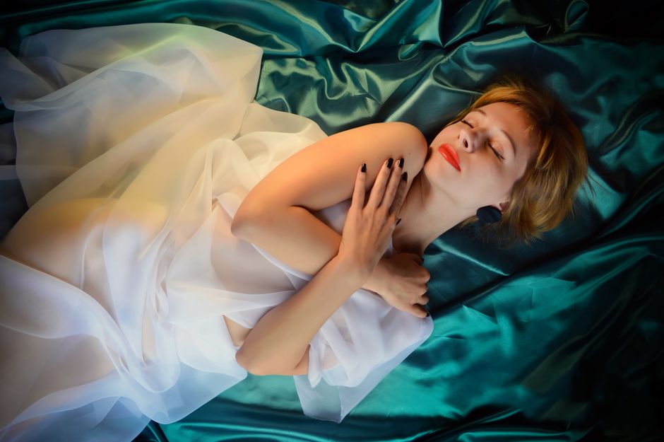 From above of young sensitive female with closed eyes and crossed arms lying on crumpled cloth