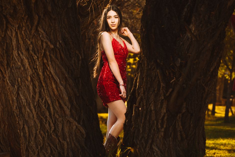 A woman in a red dress poses between trees outdoors, wearing cowboy boots.