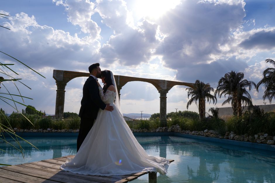 Bride and groom share a romantic kiss by a historic aqueduct under a dramatic sky.