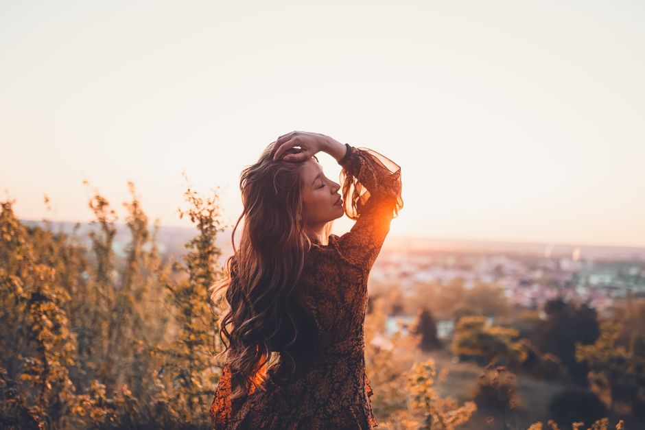 Back view of young serene female with closed eyes and wavy hair standing in grass at sunset