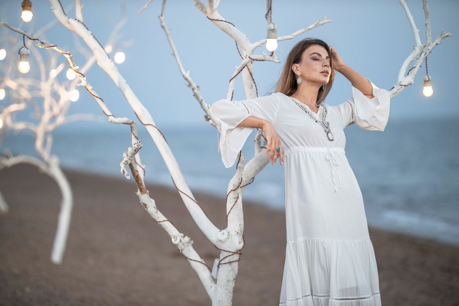 A fashionable woman posing elegantly by the beach with string lights and white branches at dusk.
