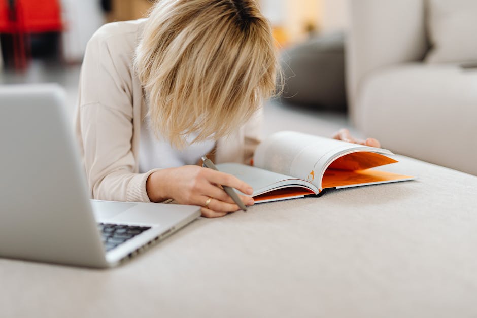 A woman reads a magazine while lying on a couch with a laptop nearby indoors.