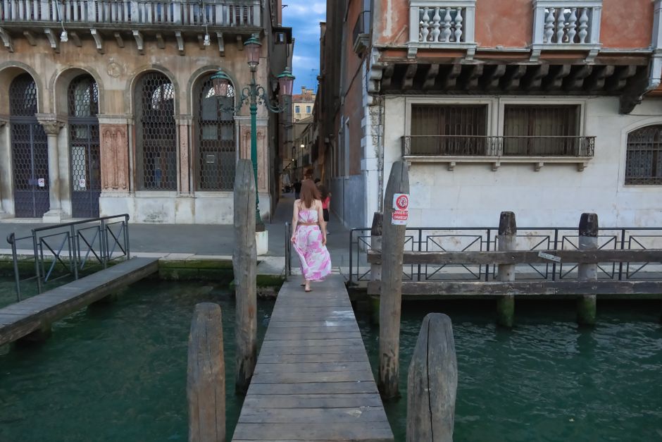 Woman in floral dress walking along a wooden dock beside a Venetian canal.