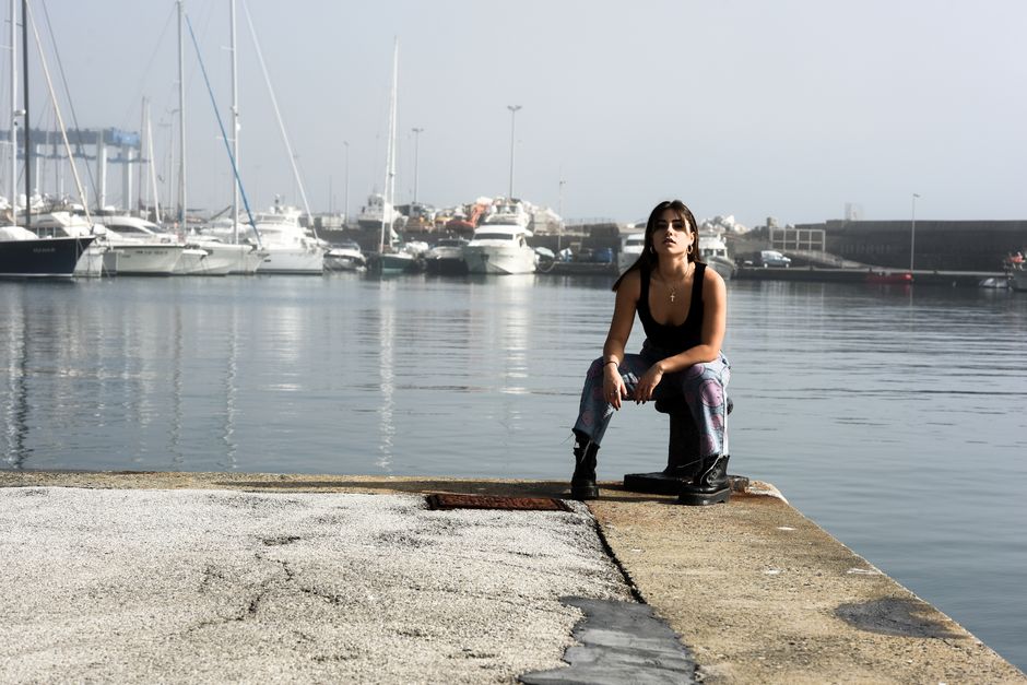 A woman in a black tank top and colorful jeans sitting on a pier with boats in the background.
