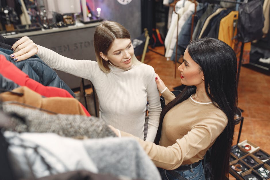 From above of young casually dressed women choosing warm puffer jacket in shop while standing near rails with clothes and discussing details