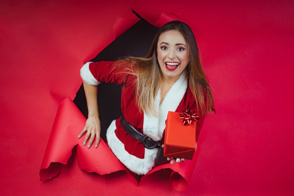 Joyful woman in Santa outfit with gift through red paper, celebrating Christmas.