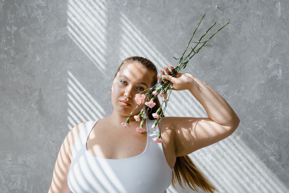 Young woman poses artistically holding pink flowers against a textured wall with natural light patterns.