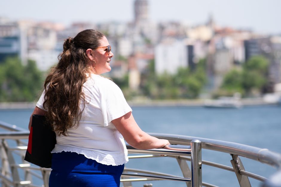 A woman stands by a waterfront railing, enjoying the scenic cityscape view on a sunny day.