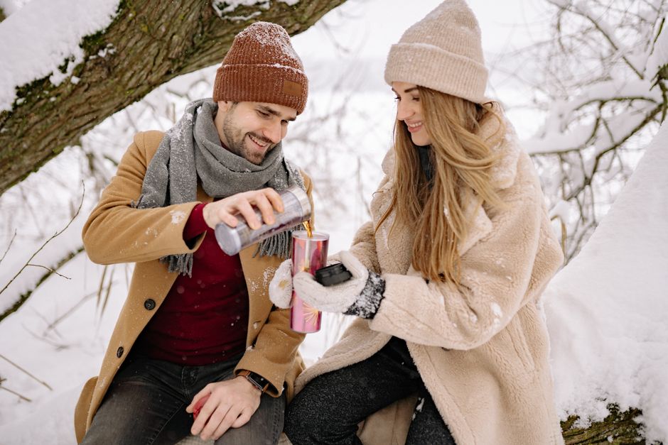 Couple sharing hot drinks under a snow-covered tree, dressed in cozy winter clothing.